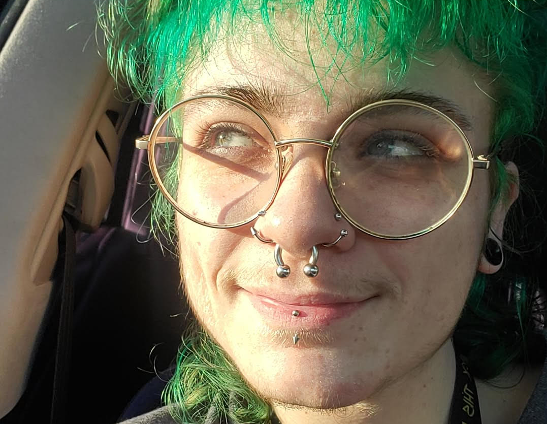 Image of a smiling person with a green mullet, round glasses, and piercings with the sun hitting their face.